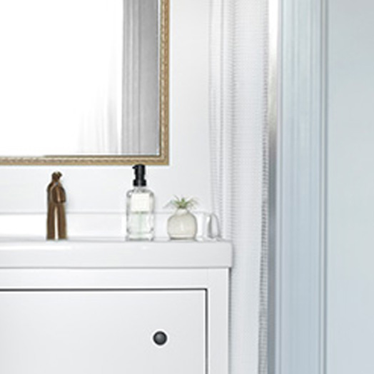 A porcelain sink with white cabinets and bronze hardware. A soap dispenser and a small succulent sit on top of the sink, along with a bronze-framed mirror above the sink. To the right of the sink is a curtain to for the shower and a light-blue painted wall.