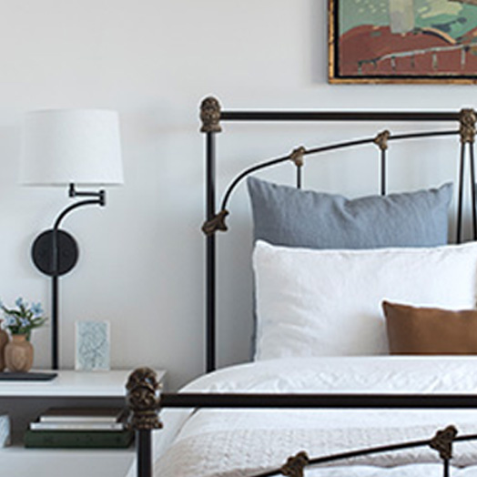 A bed with a metal bedframe with wood accents. A blue pillow, white pillow and brown pillow sit on top of the white comforter. A side table sits to the left of the bed with a lamp and two plants.